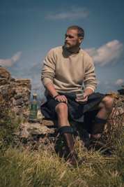 Sam Heughan sitting in Scottish landscape with glass of whiskey