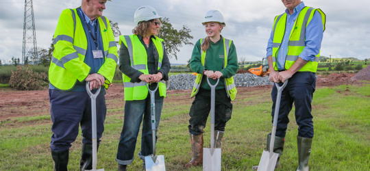 Group of people marking the start of a construction project while holding spades at a site surrounded by earthworks and construction activity.