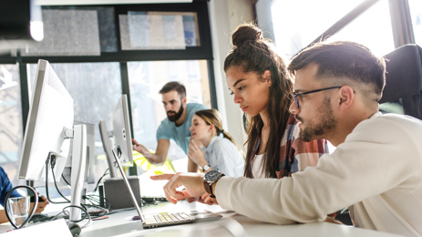 A group of people working together in a bright, modern office. Two colleagues sit at a desk in the foreground, collaborating while looking at a laptop and pointing at the screen.